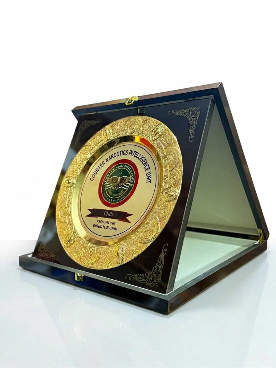 A round gold-toned commemorative plaque with an embossed border and central emblem, mounted inside a dark triangular acrylic display case on a white reflective surface.