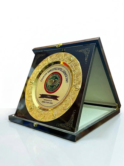 A round gold-toned commemorative plaque with an embossed border and central emblem, mounted inside a dark triangular acrylic display case on a white reflective surface.