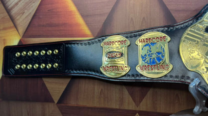 A black leather wrestling championship belt with ornate gold-tone metal plates, displayed on a geometric wood-patterned surface. The belt features two prominent gold plates with 'HARDCORE WRESTLING' text and a blue and gold central emblem.