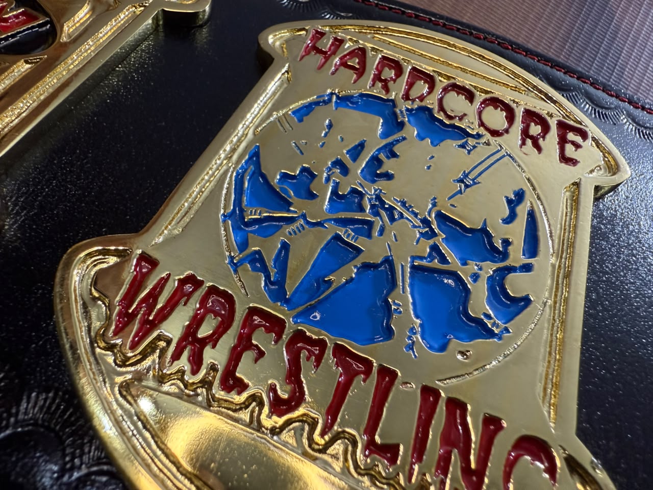 Close-up of a gold metal badge with red and blue enamel details reading 'HARDCORE WRESTLING' on a dark textured background.