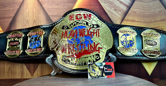 A black leather wrestling championship belt with large gold-toned metal plates, displayed on a stand against a wood-patterned background. The center plate reads 'ECW WORLDs Heavy Weight Wrestling Championship' with red and blue lettering, and the side plates are also gold-toned with multicolored details.