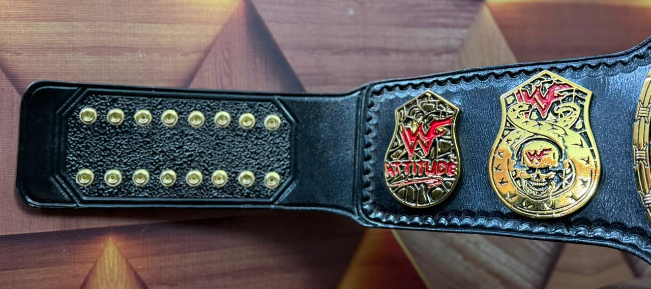 A black leather WWE Championship belt laid flat on a geometric wooden surface, showing gold-tone hardware, studs, and two championship emblems including the WWE logo.