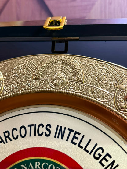 Close-up of an ornate round plaque or medallion with a decorative gold-toned border and part of the text 'NARCOTICS INTELLIGEN' visible on a white background.